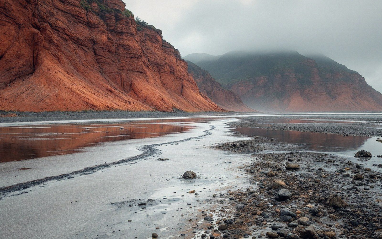 A tidal flat at low water with the high-tide line clearly marked on the red cliff behind, wide, overcast